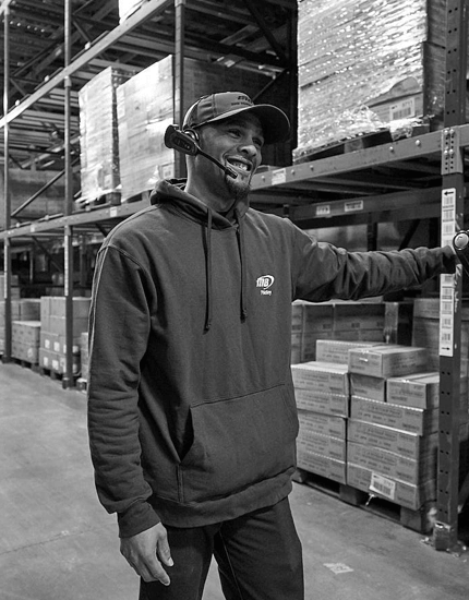 A reyes Holdings team member wearing a 2-way radio headset and standing in front of warehouse racking.